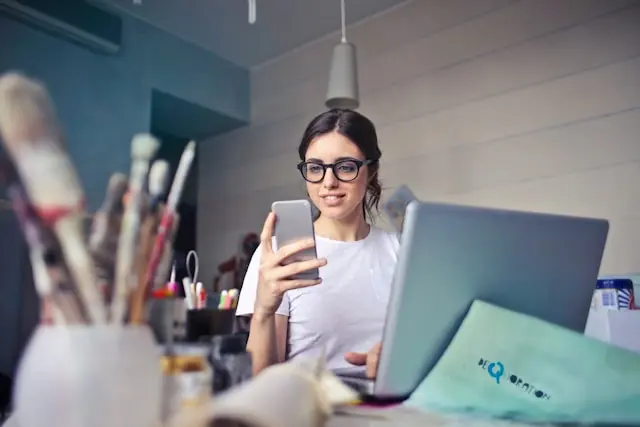 mujer frente a una laptop viendo su telefono
